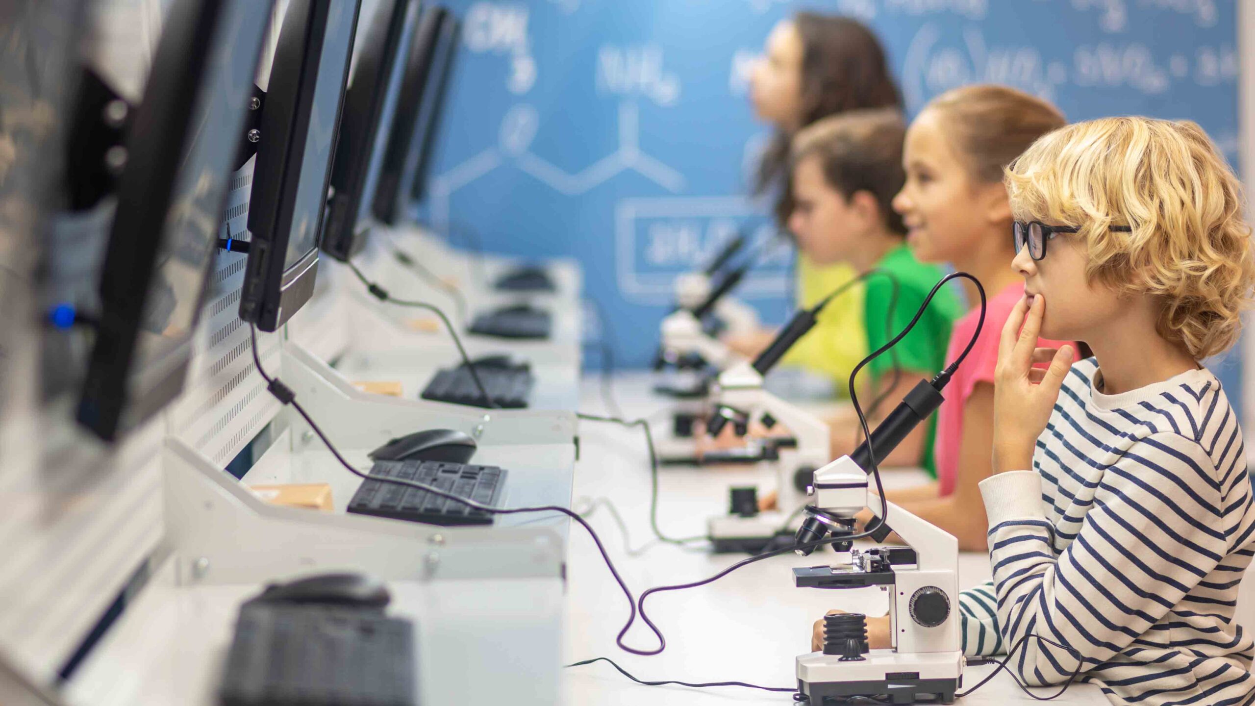 Information. Boy with glasses sideways to camera thinking looking at charging board and classmates in chemistry class